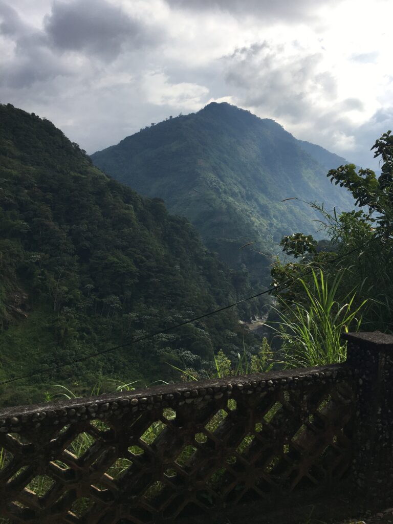Pictured is a nature scene from Baños. Pictured are two very close mountains with lots of greenery. There is also some fog present, along with a cloudy grey sky. 