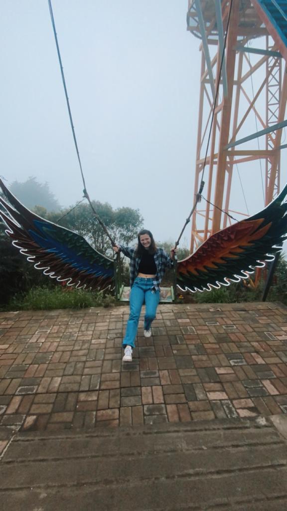 Pictured is me sitting on a swing with colorful angel wings. Behind me is a far-away background of Baños, as I am near a mountain ledge. 