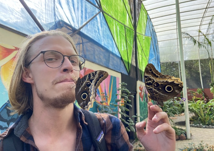 Pictured is my friend Dalton with two moth-like butterflies. One butterfly is sitting on his finger tips while the other is sitting on his face near his lips. 
