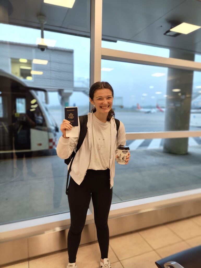 Pictured is me waiting in the airport to board my flight. In the photo, I am smiling, holding up my passport and a much-needed coffee. 