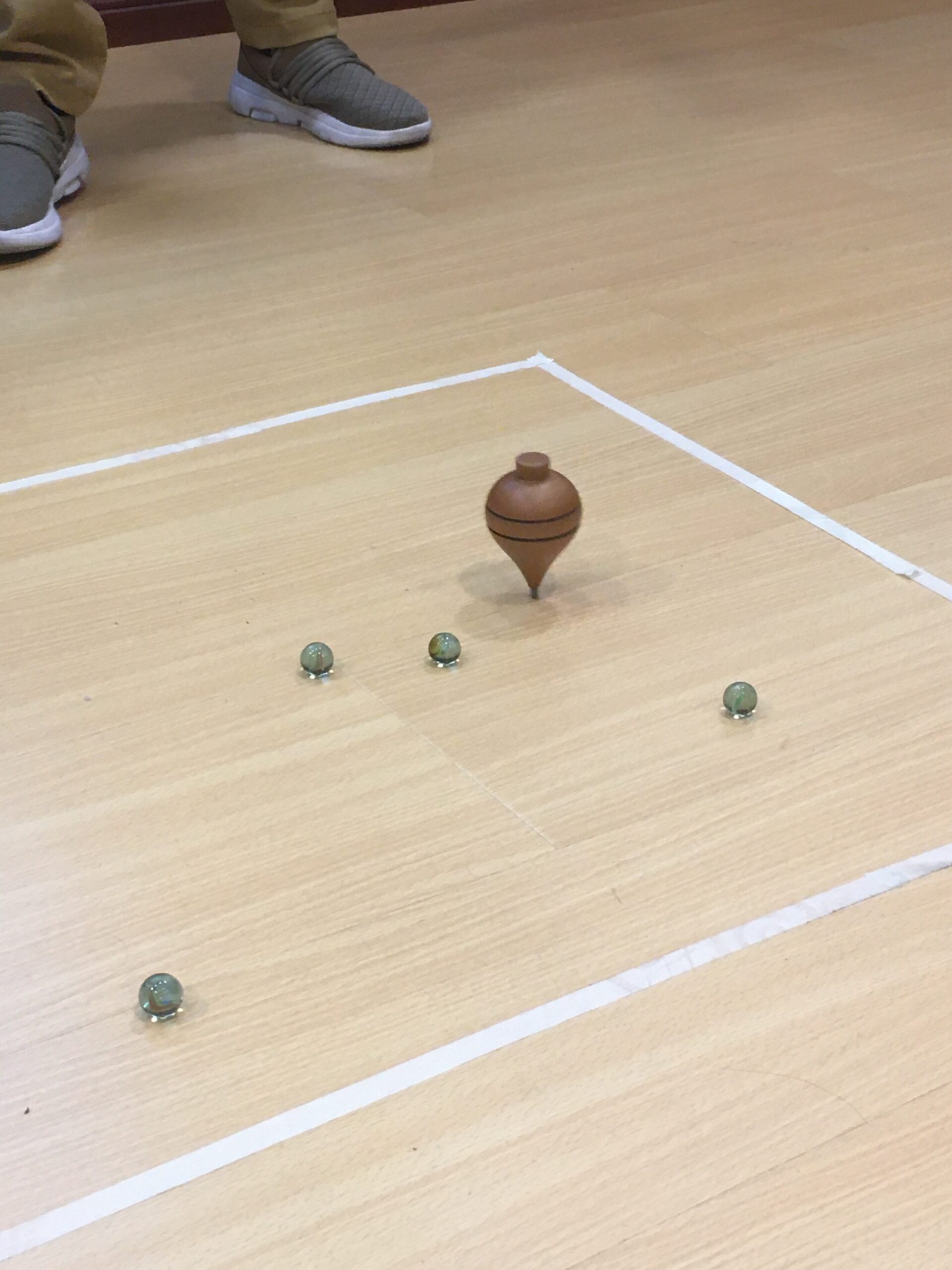 Pictured here are marbles and a wooden toy spinning on the floor. both the marbles and wooden toy and within a square, which was made by white tape. 