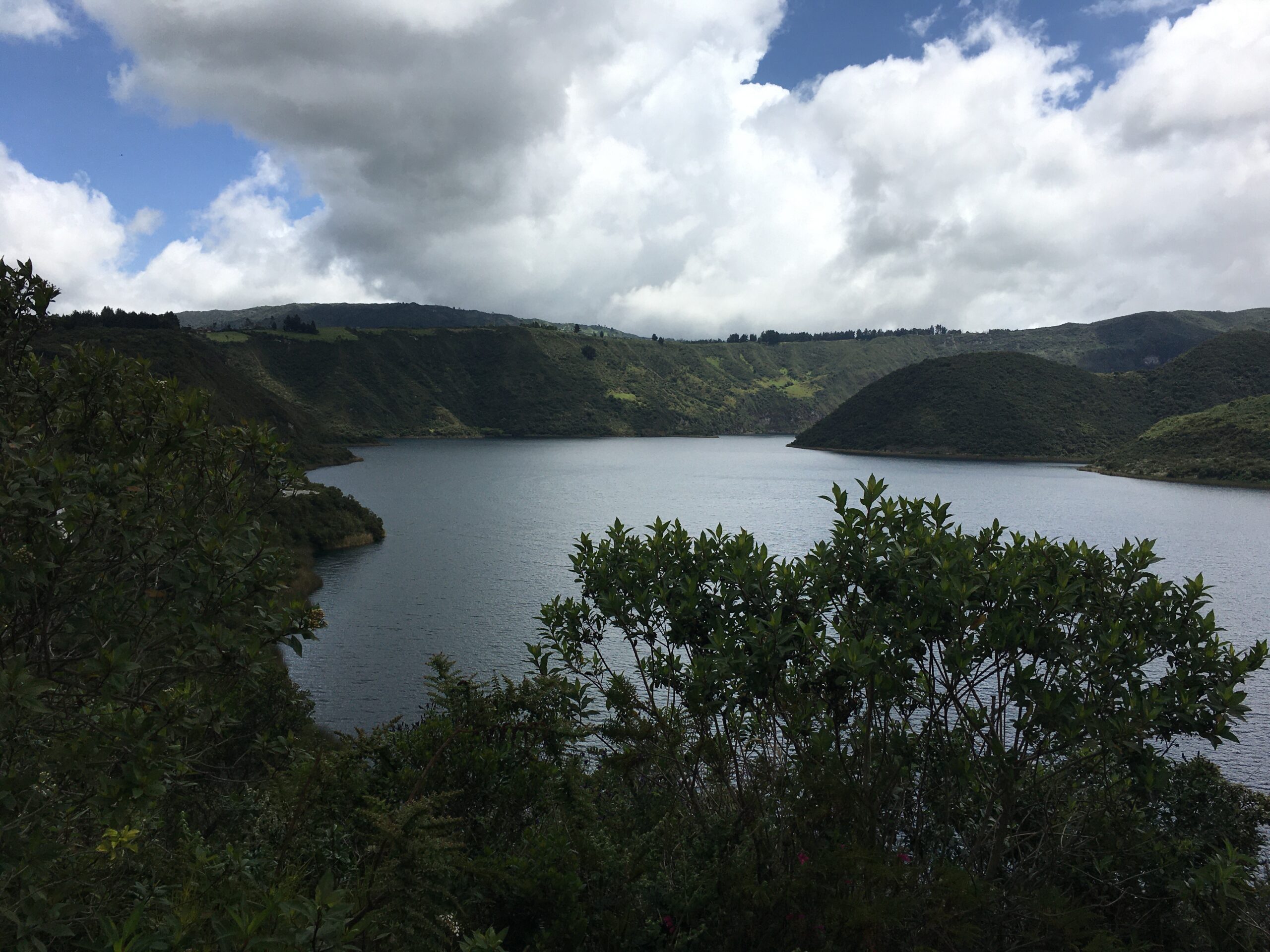 Pictured is the Cuicocha lake. The greyish-blue lake centers the photo, with green mountains surrounding the lake. 