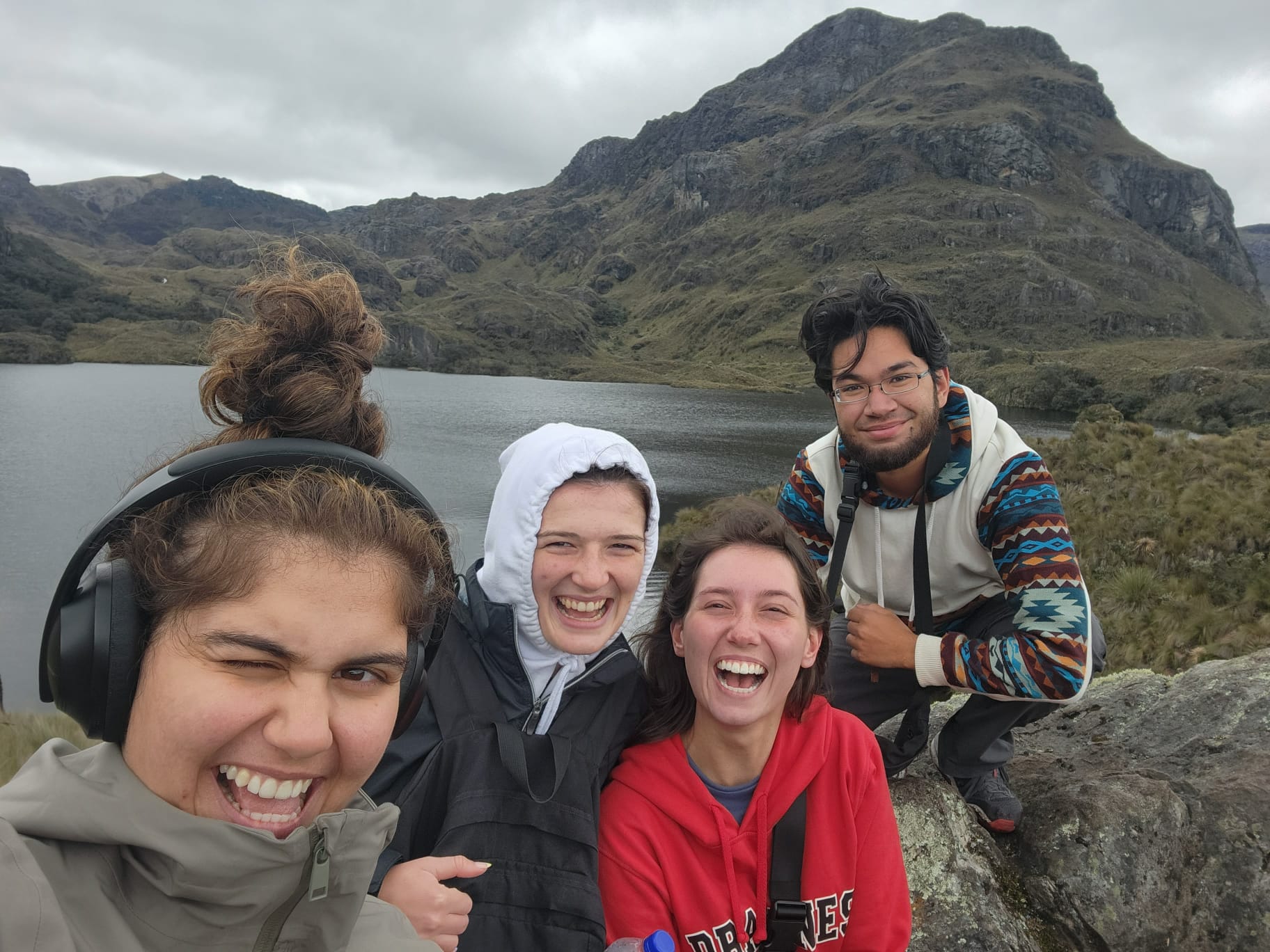 Pictured are my friends and I taking a selfie. Behind us in a lake in cajas, which is surrounded by mountainous rock and green shrubbery. 
