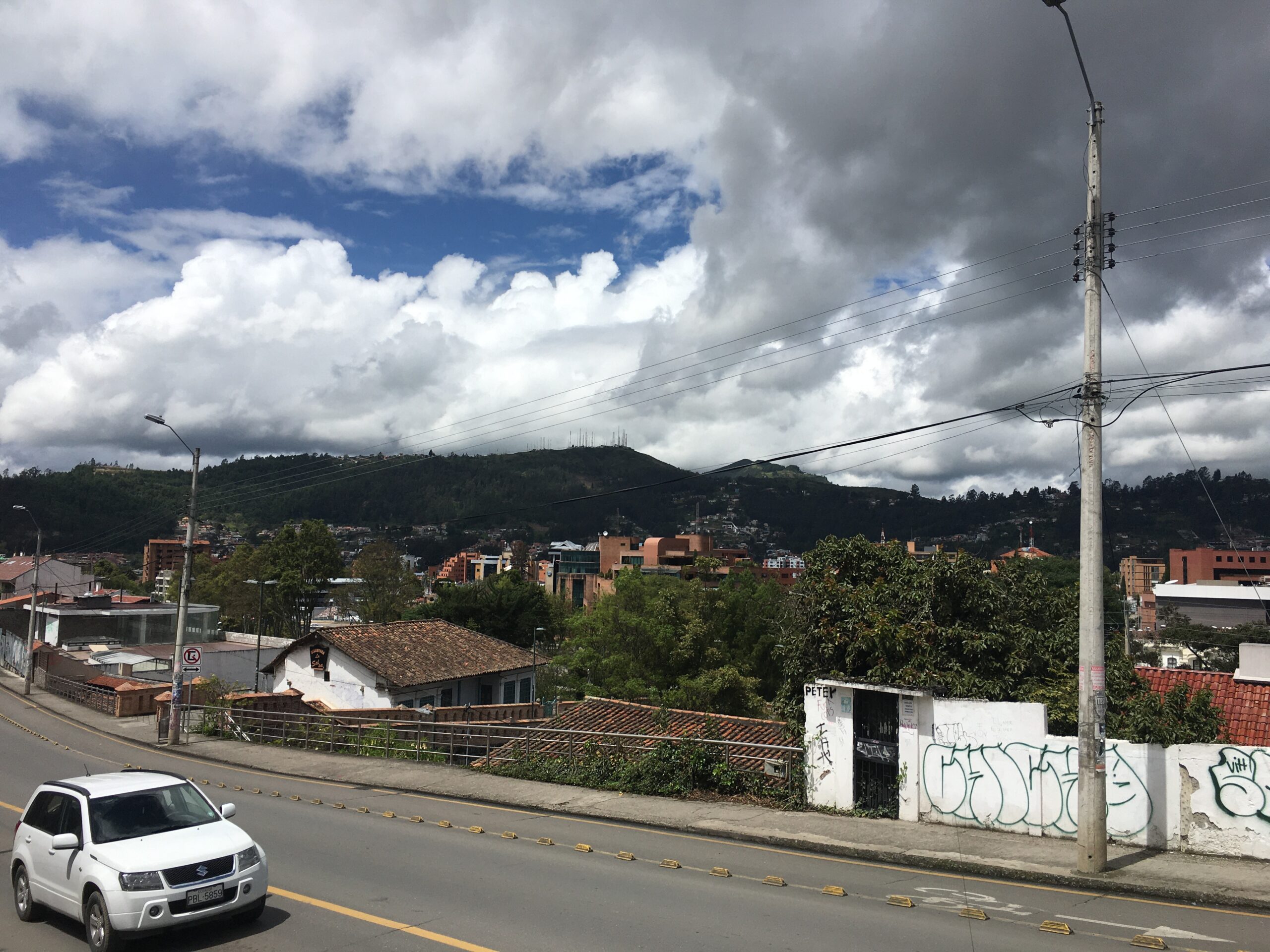 Pictured is part of the city of Cuenca. Present in the picture is a road, which is surrounded by hills and greenery, topped with a cloudy, but somewhat blue, sky. 