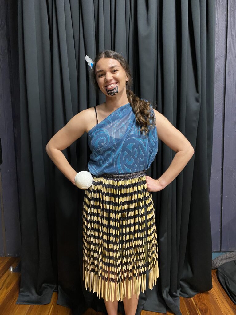 Kahiau wearing a traditional piupiu skirt and Māori scarf with the poi at the waist and a feather in her hair. 