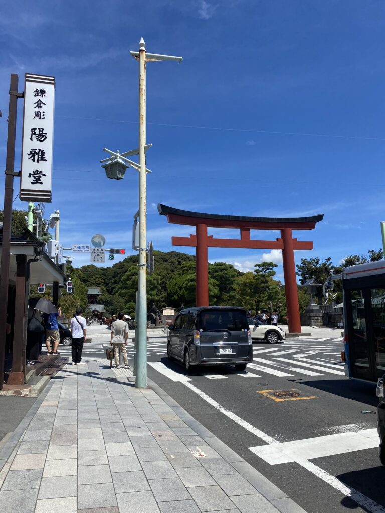 A road, there is a sign on the left side and a pole on the sidewalk near the center. On the right there is a red torii gate, which indicates the entrance to a Shinto Shrine