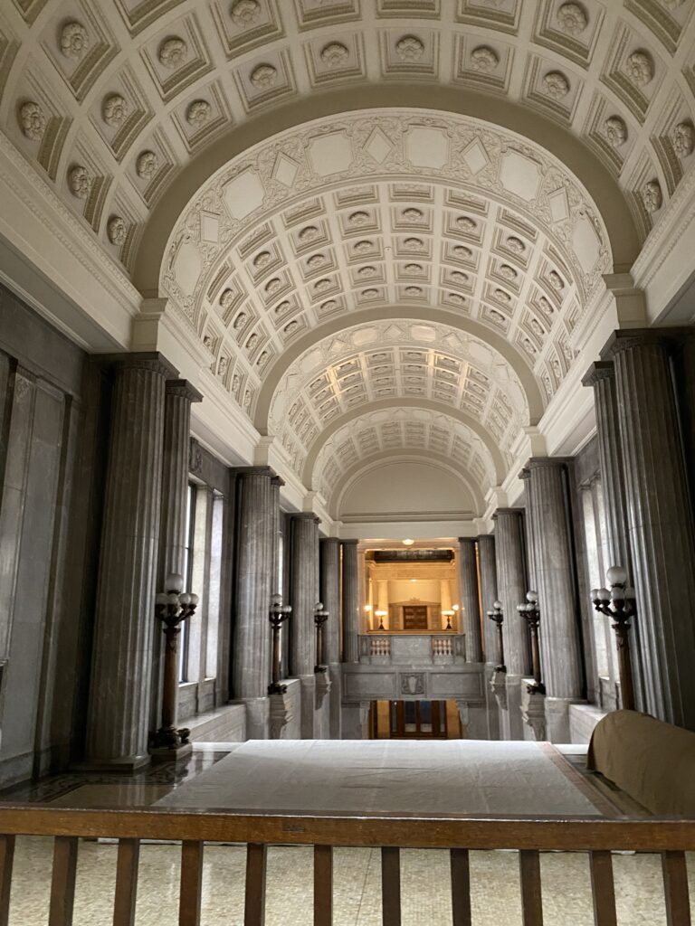 A look into another hall, the highlight of this image is the detailed ceiling. It is a white, arched ceiling with squares and flowers carved into it. There are dark gray pillars along the walls.