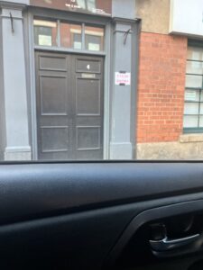 Looking out the window from an Uber to an old brick building with a set of old wood double doors, the right one of which has the number 4 in the upper middle. A sign to the right of the doors reads 10 pound entry