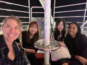 Clara, Aki, Chloe, and Abhita sitting in a circular Ferris Wheel car high above the lights of the Goose Fair shining at night below