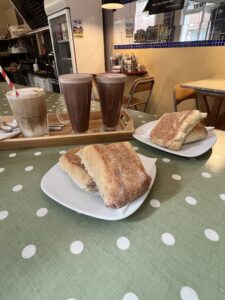 Two sandwiches sitting on two plates in the foreground, one iced latte with a spiral colored paper straw and two hot chocolates sitting on a tray in the background on a polka dotted table