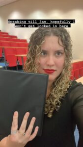 Close up of Clara, slightly smiling with curly hair flowing around her shoulders and bright red lipstick. In her hand she holds a small black binder, and behind her are rows of red fabric seats and desks