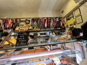 An order counter at a Florentine sandwich shop with breads and meats in the foreground and cooktops behind the counter with lots of penents and framed photos lining the wall behind it