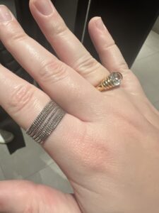 Clara’s hand, with a ring on her pointer and ring fingers. The ring on her pointer finger is 7 very thin rows of silver beads all in line with one another to make a very thick band. The ring on the ring finger is a gold band that gets wider at the top and sets a clear, fake diamond