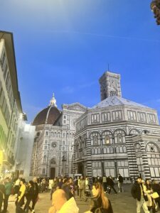 An alleyway alongside the Duomo showing the main entrance as well as part of the side that shows the dome peeking out in the background. The photo was taken at sunset, so the sky is a bit darker blue and the warm streetlight gives the whole building a very warm, inviting glow
