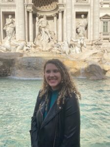 Clara in the foreground standing in front of the Trevi Fountain with a large smile on her face