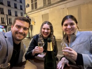 Olin, Clara, and Shila sitting from left to right in a selfie. Olin is taking the selfie and Clara and Shila are holding wine glasses with champagne in them. The bottle of champagne sits on the table in front of them and the street lies behind them at dusk