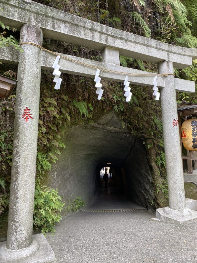 Cement Torii gate that is not colored red with a rope with white thunderbolts strung across it. The entrance to the shrine appears to be a cave-like tunnel.