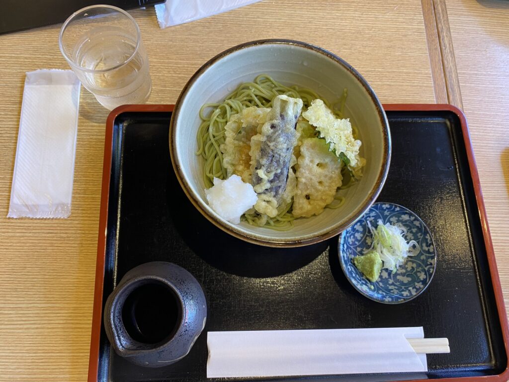 A black lunch tray with a bowl of the broth for the soba noodles, a small plate with cut green onions and wasabi, with the main bowl having matcha soba noodles topped with vegetable tempura.