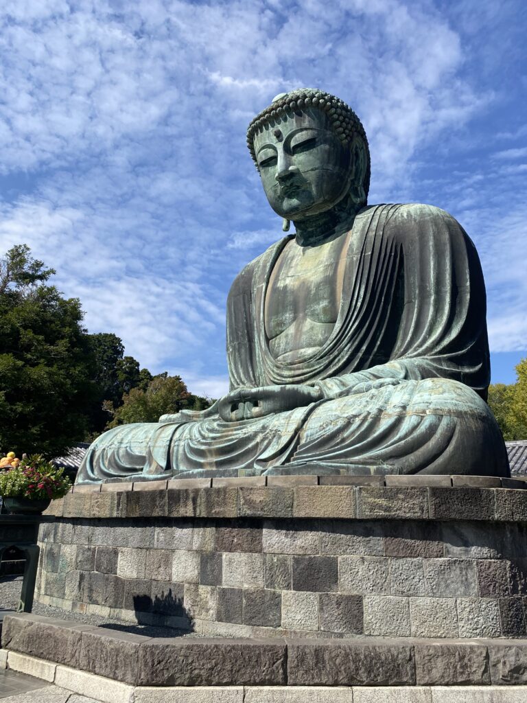 A large statue of Buddha, it is an oxidized shade of green with some darker areas. The state is sitting on a base of stone bricks.
