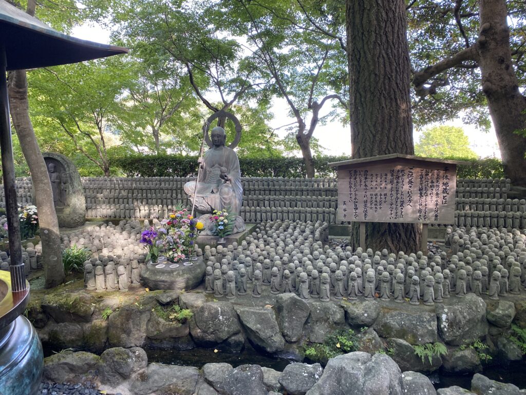 To the left of the center, there is a medium size statue that is surrounded by many small jizo statues. The bigger statue has flowers placed in front of it.