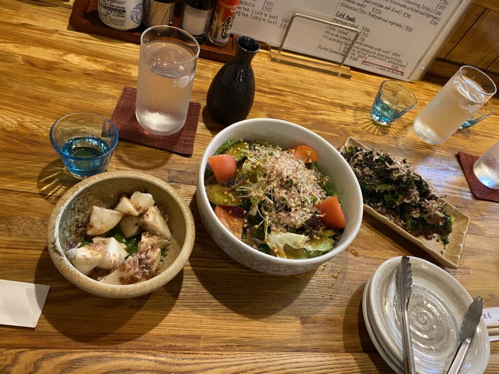 A very small array of dishes. On the left is small bowl of mountain yams with bonito flakes and other seasonings. In the center is a bowl of a garden salad. On the right is a plate with sliced cucumber topped with bonito flakes and other seasonings. 