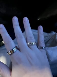 Clara’s hand, outstretched, showcasing 3 rings. A silver band with two engraved lines on the pointer finger, a silver and gold indented diamond pattern and edging on the ring finger, and a silver indented diamond patterned ring on the pinky finger