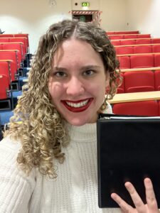 Clara, fully smiling with hair down, red lipstick, and a white turtleneck top is holding a small black binder in front of her. Behind her, you can see the same lecture theater seats