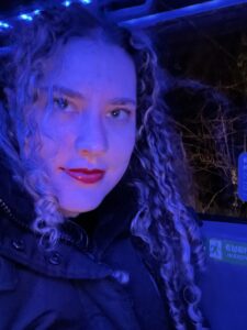 Clara with a slight smile sitting in a window coach seat. There is a bluish light to her face because of the blue coach lights