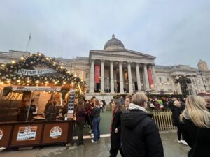 A London Christmas market. Lots of people in the foreground with some lights strung up along the stalls, and a larger building with pillars sits in the distance