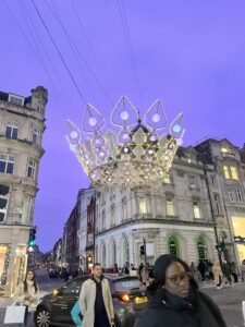 A large crown made of lights sits suspended in the air over an intersection at dusk with some buildings behind it