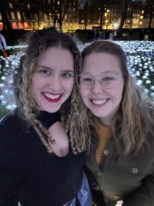 Clara (left) and Lena (right) standing in front of a field of lighted white roses smiling for a selfie