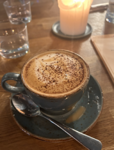 A picture of the aforementioned delicious cappuccino. Blue cup and saucer with a foamy drink inside. Dusting of cocoa powder on top.