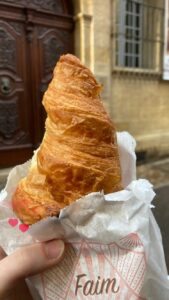 Holding a croissant in front of a building with wooden carved doors.
