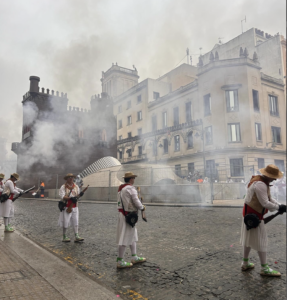 locals dressed in period wear shooting their guns into the air. Smoke billows around old architecture and a foam castle.