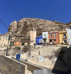 view of the neighborhood in front of the sandy cliff face and castle. One very blue house, and smaller, less colorful houses. 