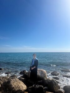 Me holding a camera in front of the view of the Mediterranean standing on large rocks.