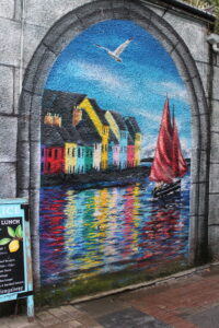 Street art of the bay in Galway. Houses along the bay are colorful and reflecting along the water. A sailboat is featured in the frame. 