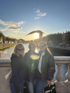 Three girls standing on a bridge during sunset, a bird is flying over head. 
