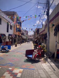 A small street in the town of Otavalo. The street is paved with colored bricks and many flags hang across the street from the rooftops above.