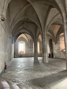 Interior view of castle, large empty room with archways supported by two center beams. Made of grey stone.