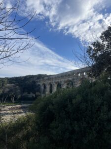 Distant view of roman aqueduct, resembles a stone bridge. Bright blue sky in the background and greenery surrounding the aqueduct.