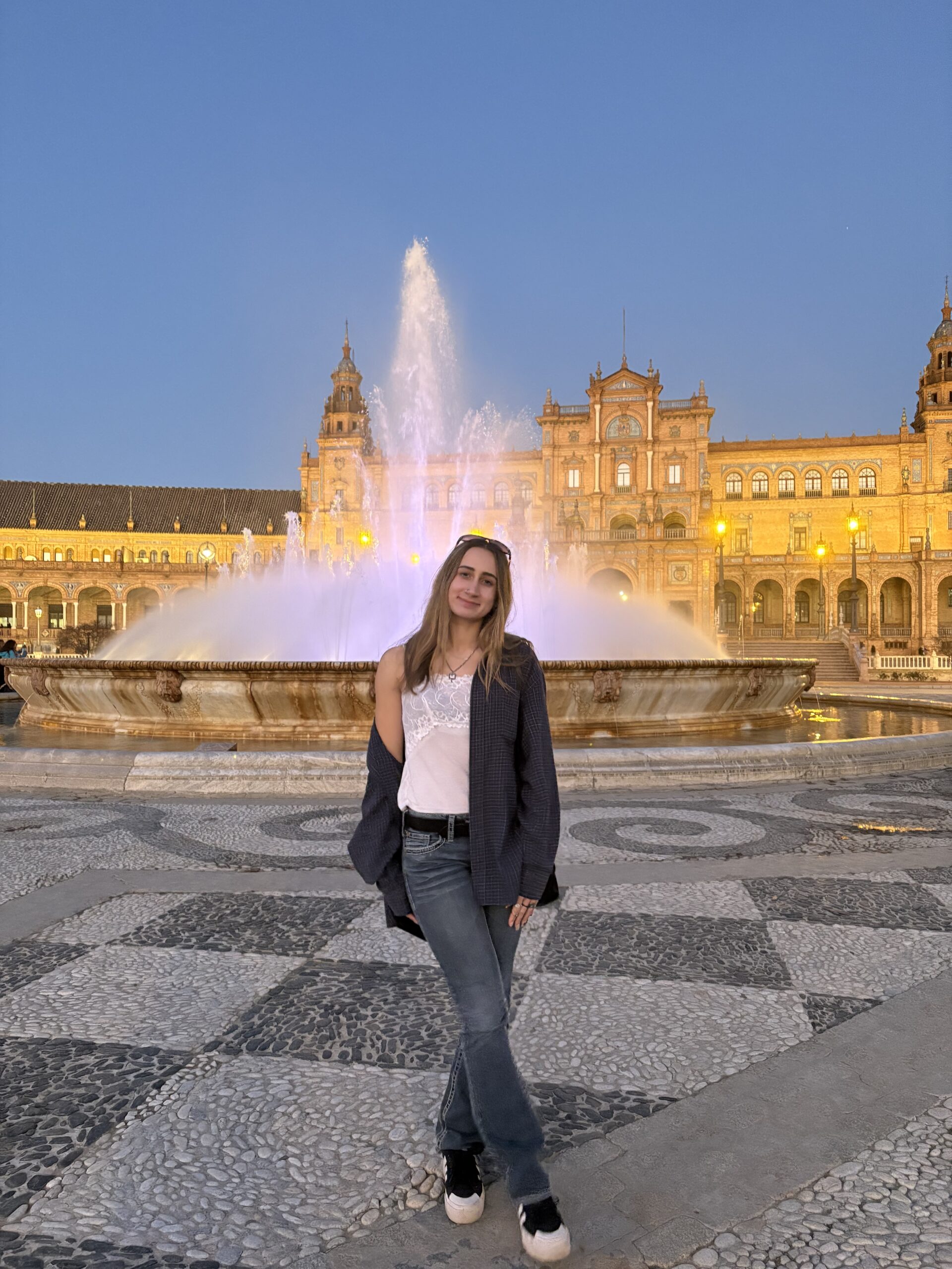 A photo of me in front of the water fountain in a big plaza called La Plaza de España, with a checkered pattern on the ground and a glowing golden building in the background