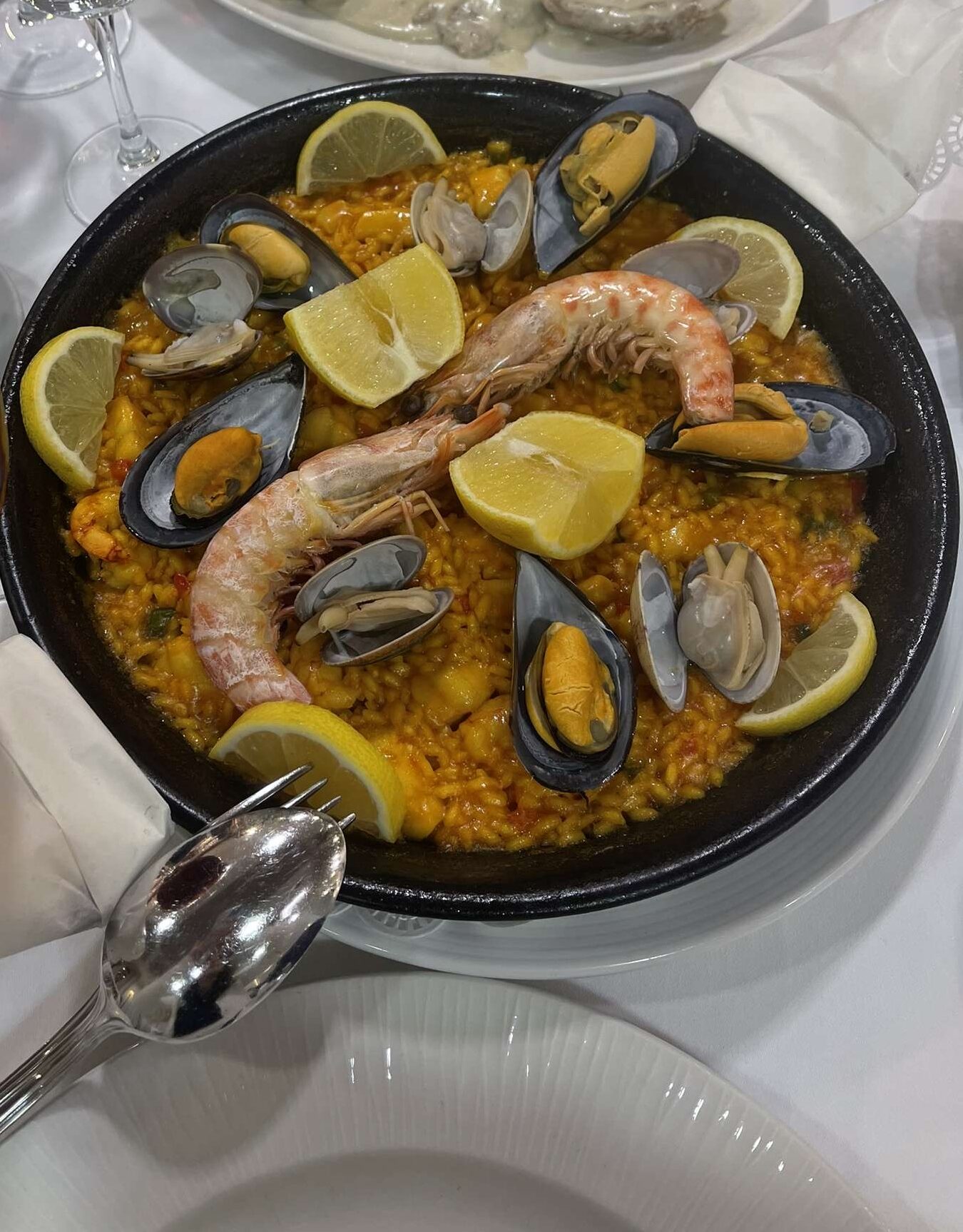 A plate of paella with shrimp, oysters, clams, lemon, and rice, displayed with silverwear