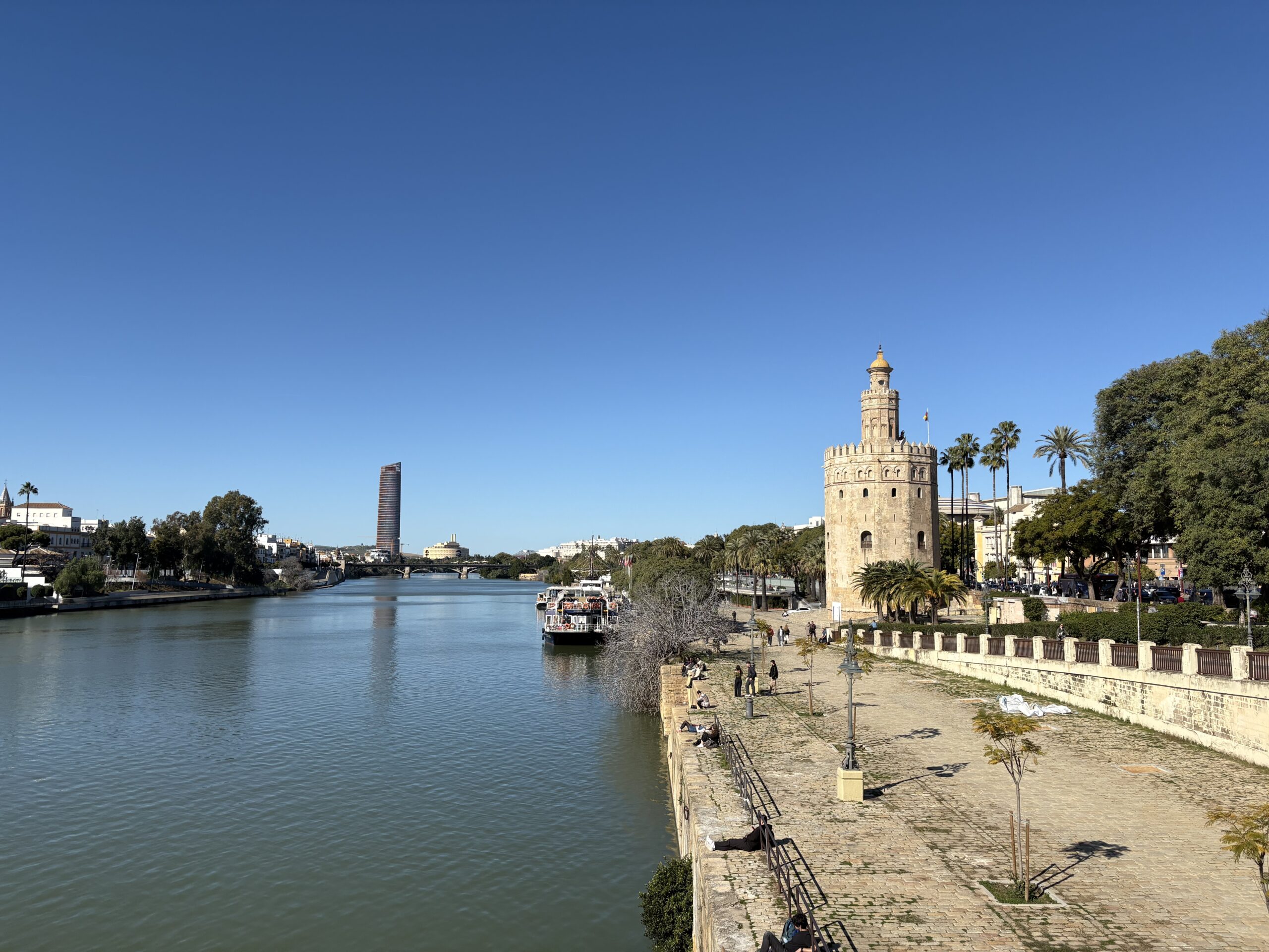A photo of a blue/green river from the view of a bridge, with a tall building to the right called La Torre de Oro, with green trees to the right of that and a pathway below next to the river