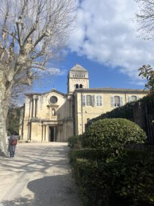 A large white building, with a circular window and a small tower to the right. Blue sky in the background with shrubbery in the foreground