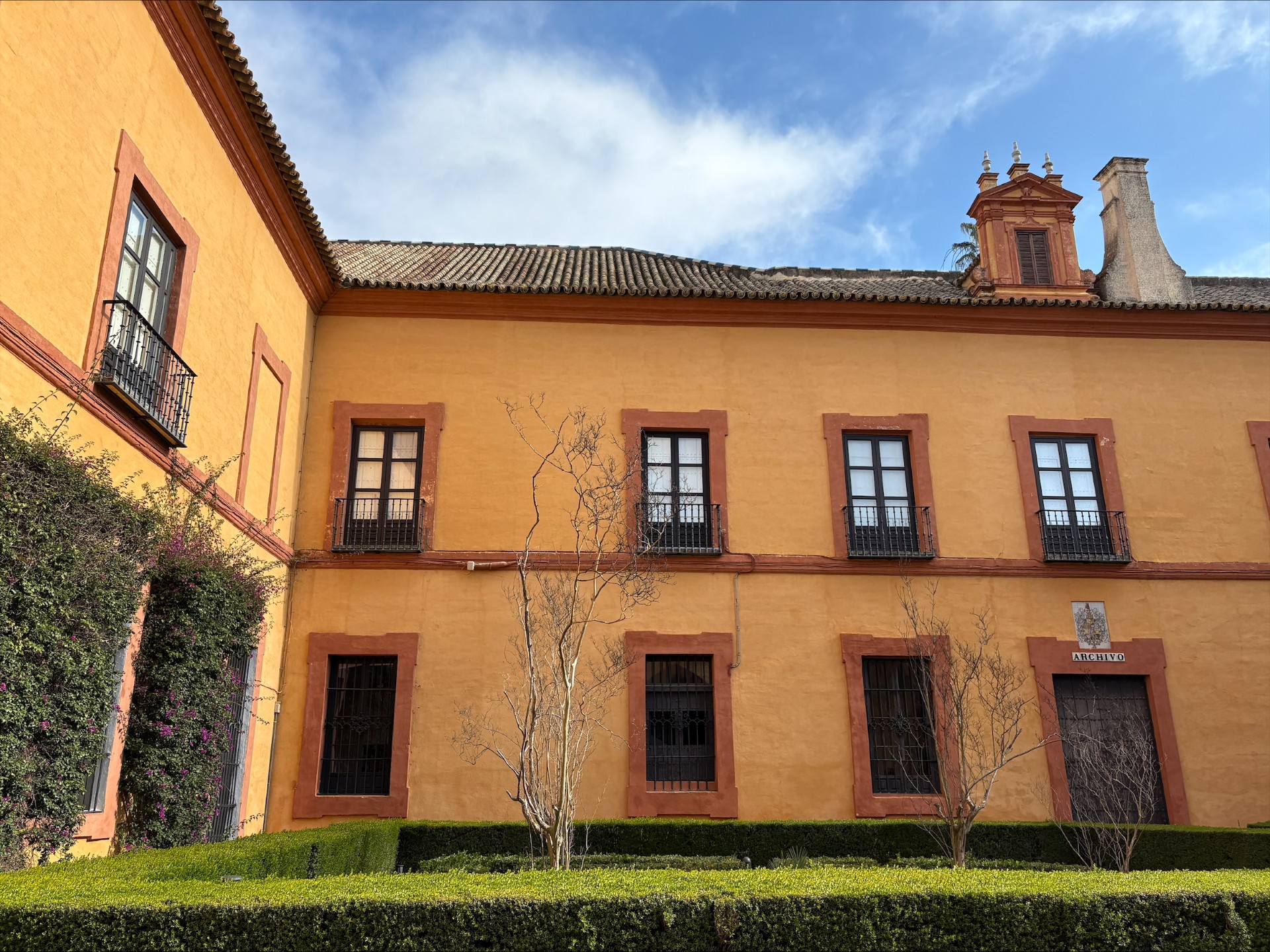 One of the buildings from the Royal Alcázar, it is a two story orange wall viewed from a garden outside, with rows of windows on both floors