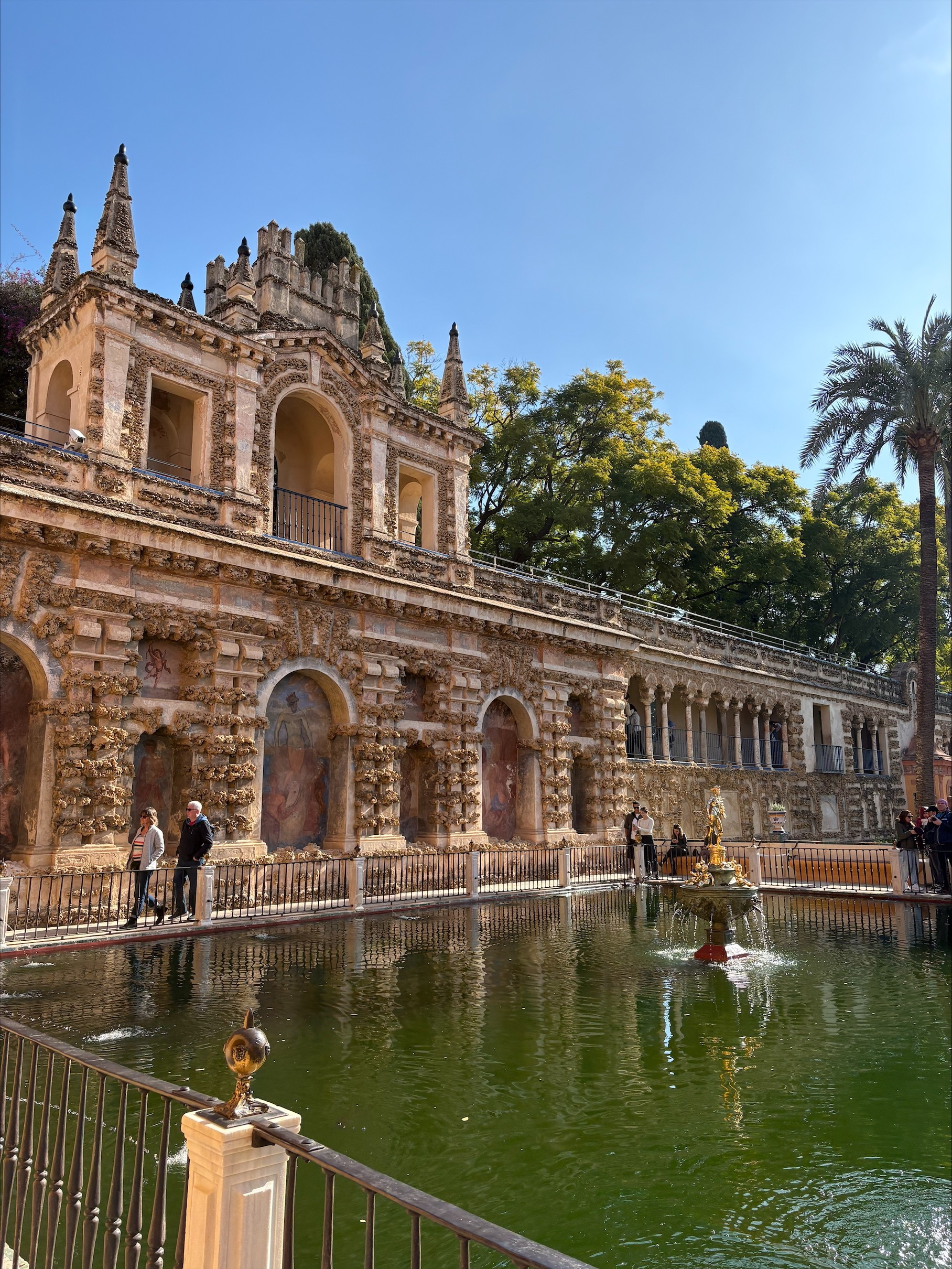 A photo of another outside garden in the Alcázar, with a fountain in the middle with green water and an orange/pale rustic colored building in the background