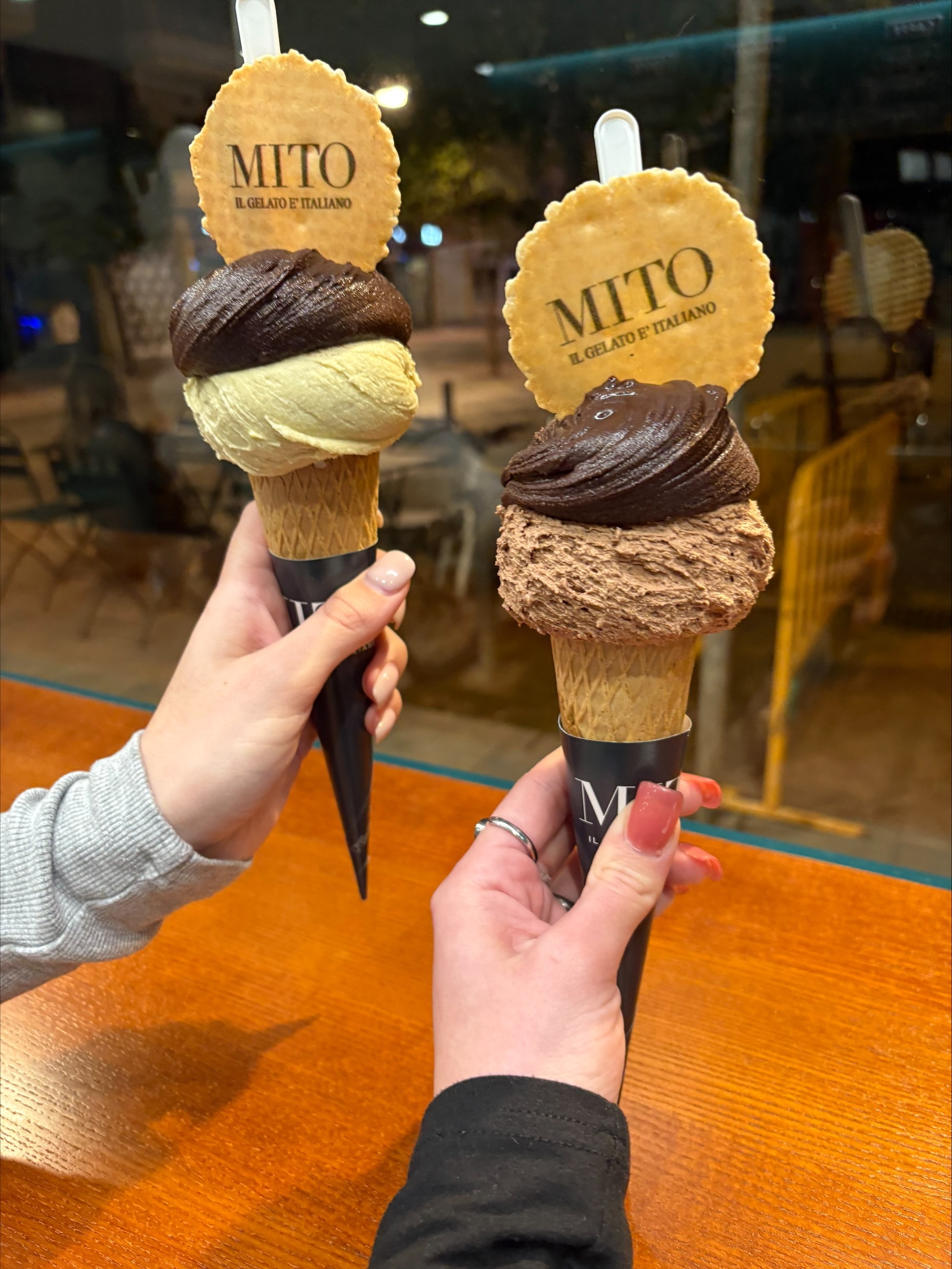 Two ice cream cones from Gelatería Mito held in front of a window, with two scoops each with cookies on top, one cone is of pistachio and dark chocolate, the other of Nutella and dark chocolate