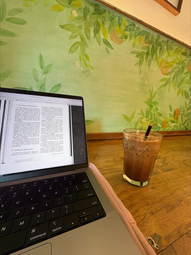 A photo of an iced coffee and my computer with homework on a brown counter at a coffee shop close to my school, with a green wall with flowers in the background