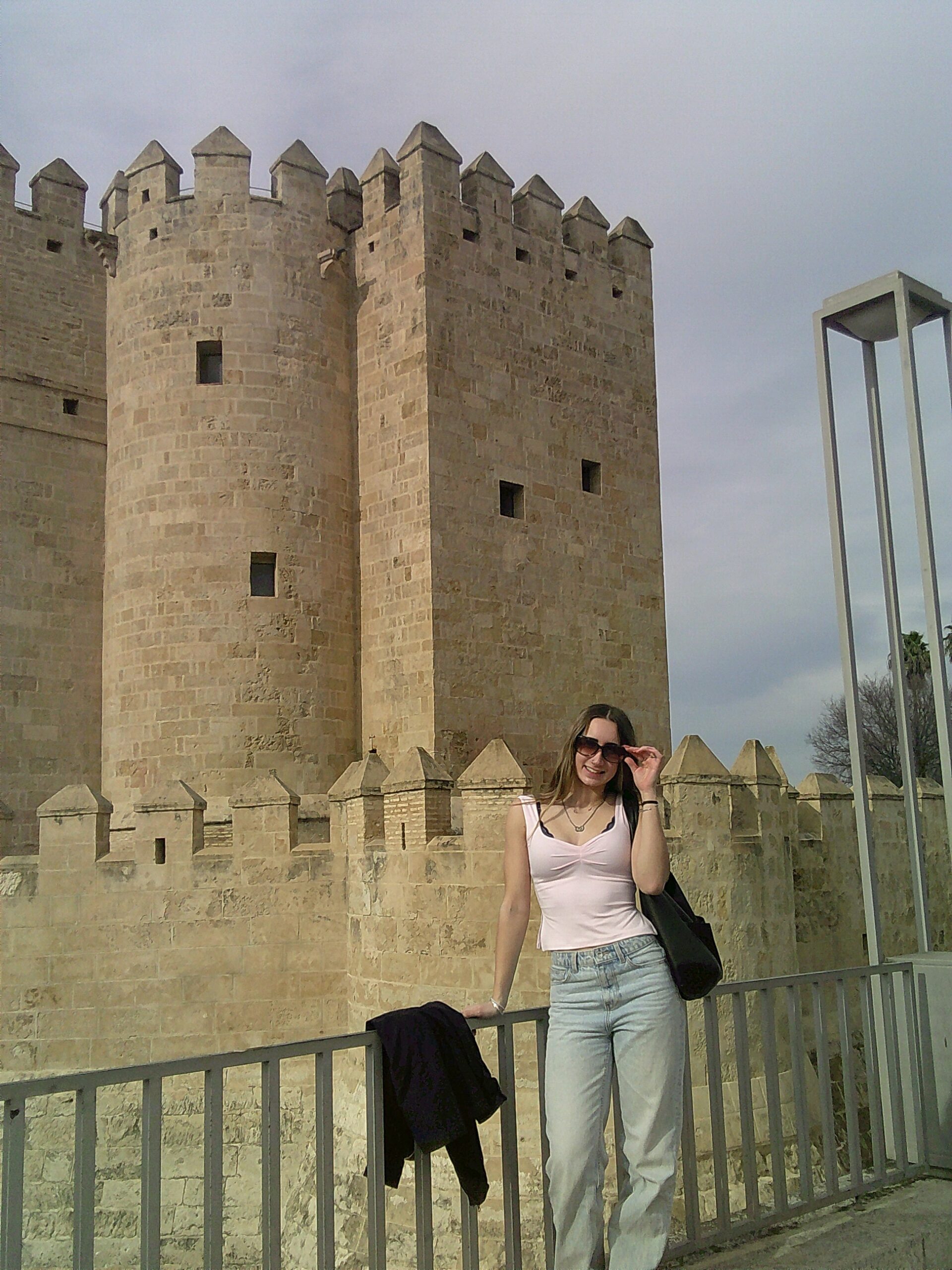 Me next to a castle in Córdoba, standing in blue jeans and a pink shirt and sunglasses leaning against a silver railing, with the beige figure over the castle rising behind me in the back